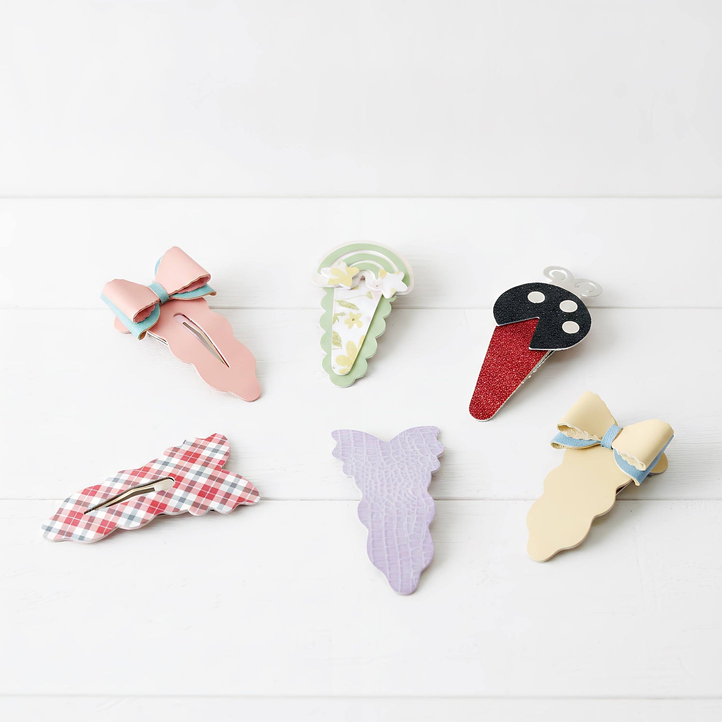 Six decorative hair claw clips arranged on a white wood background. The clips feature pastel and patterned designs including a pink bow, a green and white floral ice cream cone, a red glitter ice cream cone with a black face, a red-and-white gingham bow, a lavender mermaid tail, and a pale yellow dress-shaped clip with a blue bow.