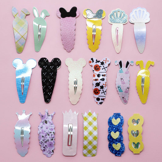 A set of 18 novelty hair clips arranged in a 3x6 grid on a solid pink background. The clips feature various whimsical shapes, patterns, and colors including plaid, glitter, floral, and cartoon motifs.