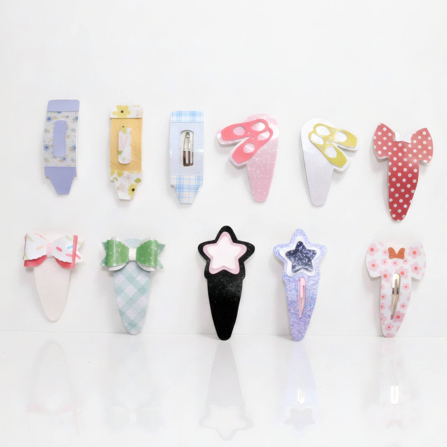 Twelve cute hair clips arranged in two rows on a white reflective surface. The clips feature various whimsical shapes including bows, stars, mushrooms, and cartoon-style heads, decorated with pastel patterns like polka dots, plaid, and florals.
