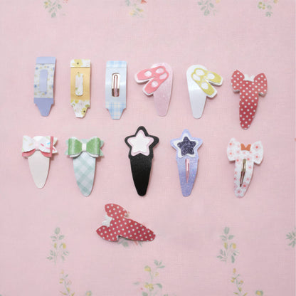 A set of ten cute, whimsical hair clips arranged on a light pink floral background. The clips feature various shapes including a tube, a mushroom, a bow, a star, and polka-dot patterns in pastel and bright colors.
