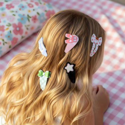 A close-up lifestyle shot of a young girl with long wavy blonde hair, viewed from behind, wearing four cute hair clips in pink, white, green, and black. She is lying on a pink and white checkered blanket next to a floral pillow.