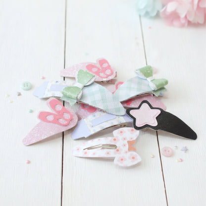 A pile of pastel paper clip hair accessories on a white wooden surface, including bunny, bow, and star designs in pink, green, blue, and black.