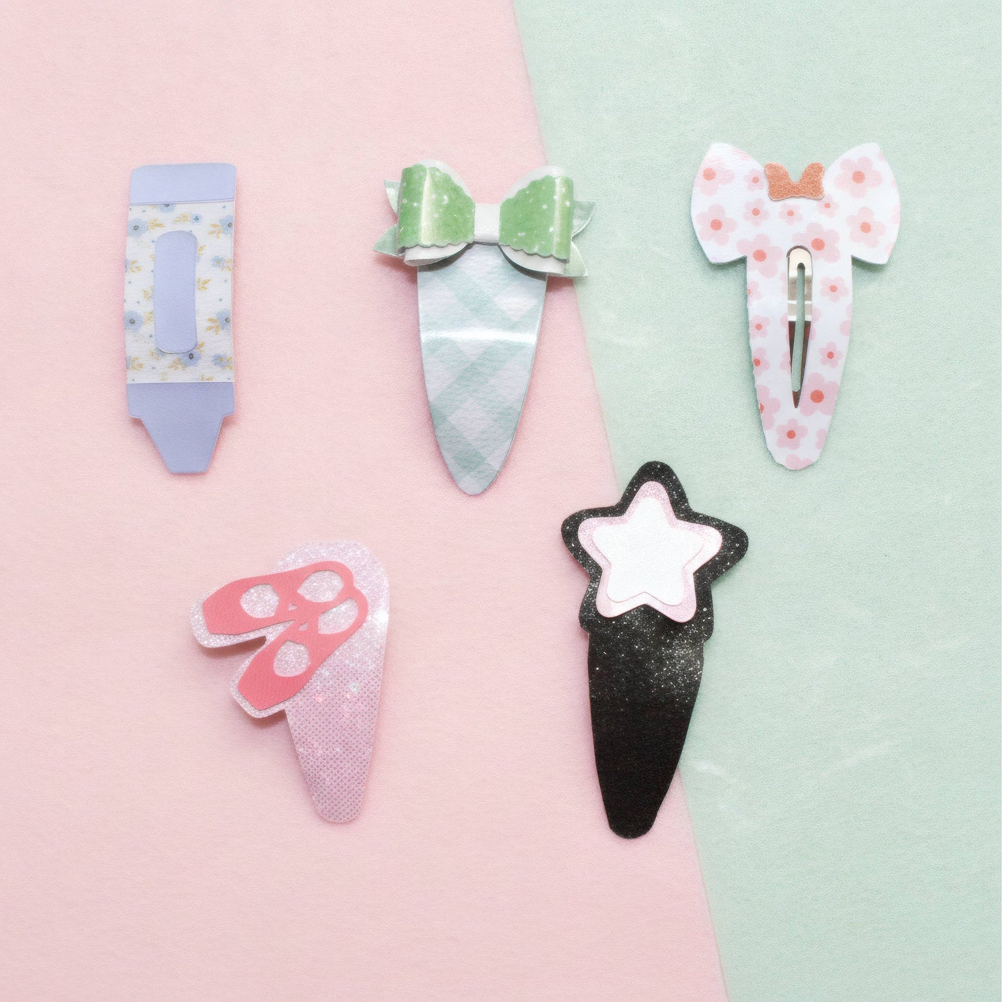 Five cute hair clips styled like sweets and accessories, arranged on a split pastel pink and mint green background. The clips include a lavender pencil, a green bow hair clip, a pink elephant ear, a pink butterfly, and a black ice cream cone with a white star.