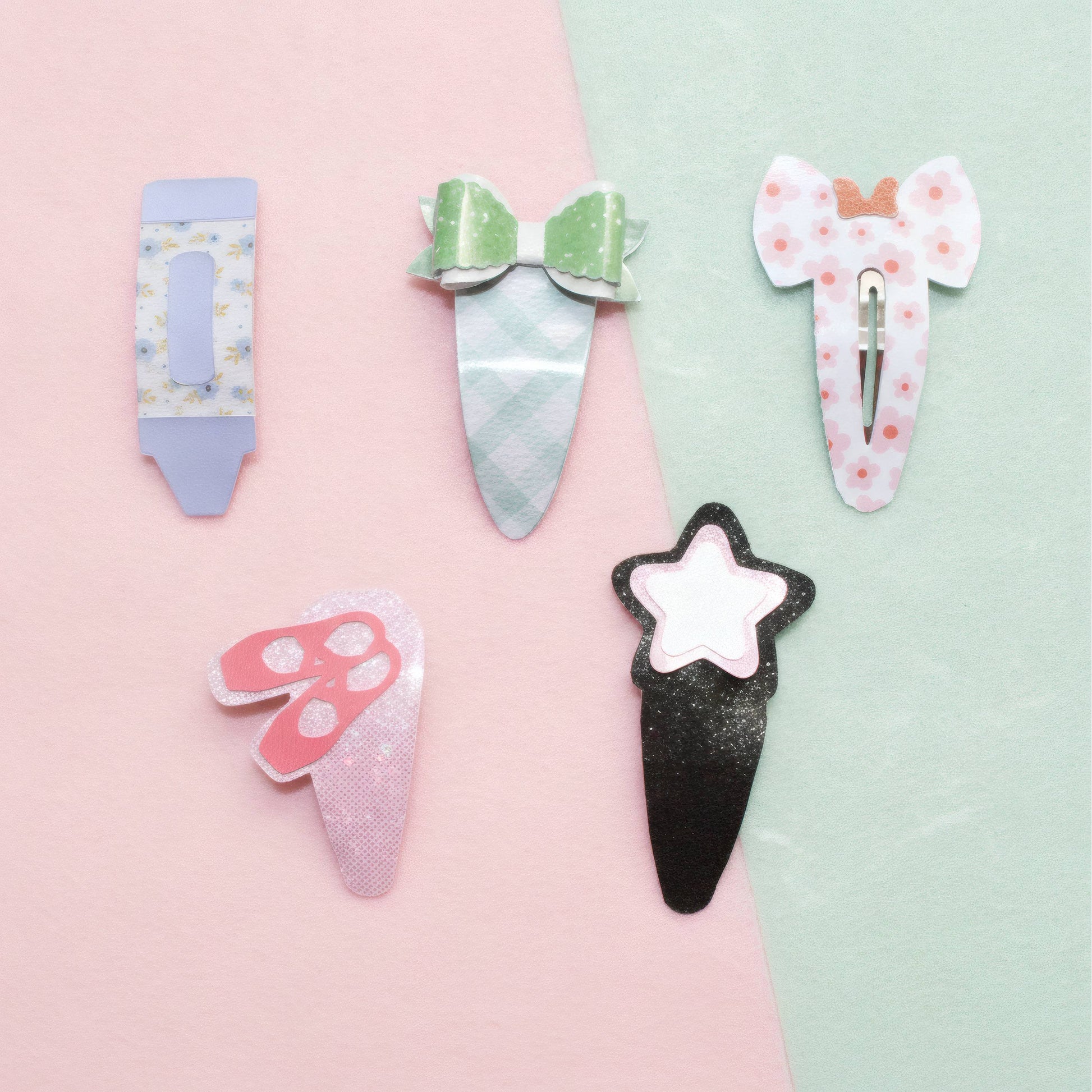 Five cute hair clips styled like sweets and accessories, arranged on a split pastel pink and mint green background. The clips include a lavender pencil, a green bow hair clip, a pink elephant ear, a pink butterfly, and a black ice cream cone with a white star.
