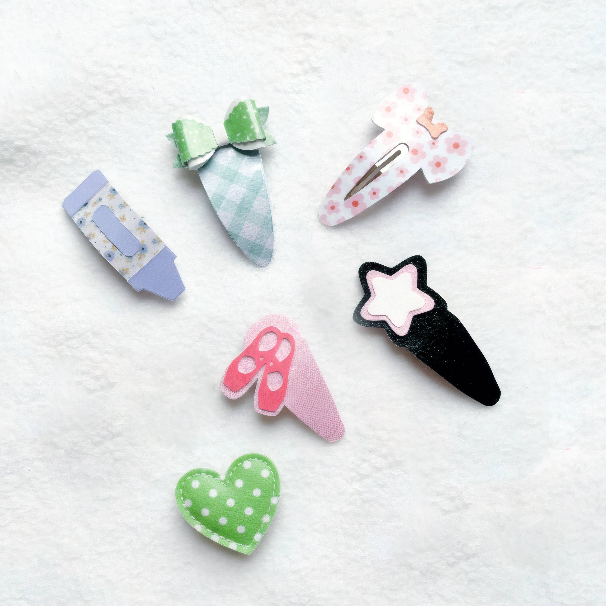 Six decorative hair claw clips arranged on a white textured fabric background. The clips feature whimsical shapes including a pencil, a carrot, a flower, a pair of scissors, a star ice cream cone, and a green polka-dot heart.