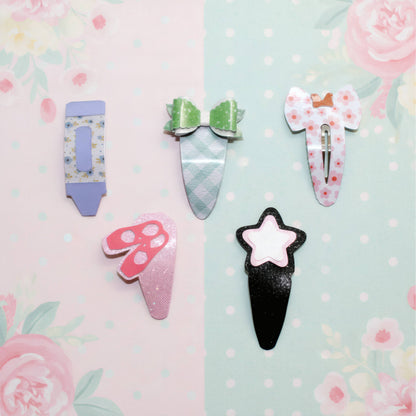 Five cute, pastel-themed hair clips arranged on a split pink and mint green floral background. The clips include a lavender tube, a green bow, a pink mushroom, a black ice cream cone, and a white bow with pink flowers.