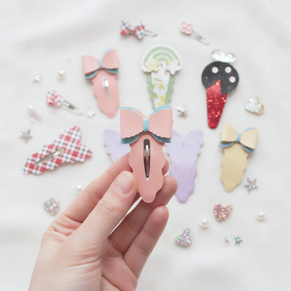 A hand holds a pink bow hair clip in the foreground, with several other pastel and patterned hair clips arranged on a white fabric background.
