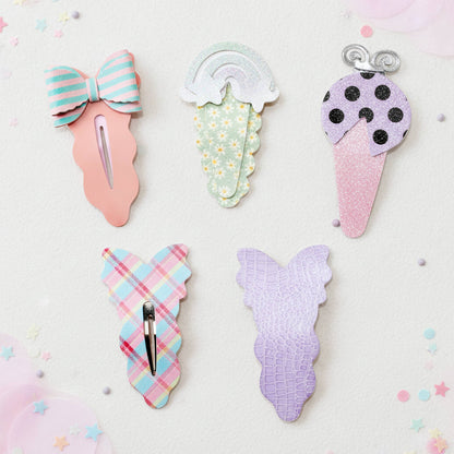 Five pastel hair claw clips styled like ice cream desserts, arranged on a light pink background with scattered pastel confetti. The clips feature pink, mint green, lavender, and plaid patterns.