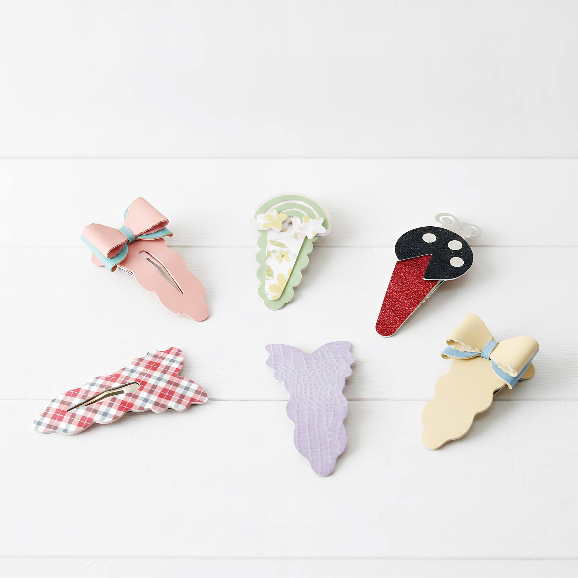 Six decorative hair claw clips arranged on a white wood background. The clips feature pastel and patterned designs including a pink bow, a green and white floral ice cream cone, a red glitter ice cream cone with a black face, a red-and-white gingham bow, a lavender mermaid tail, and a pale yellow dress-shaped clip with a blue bow.