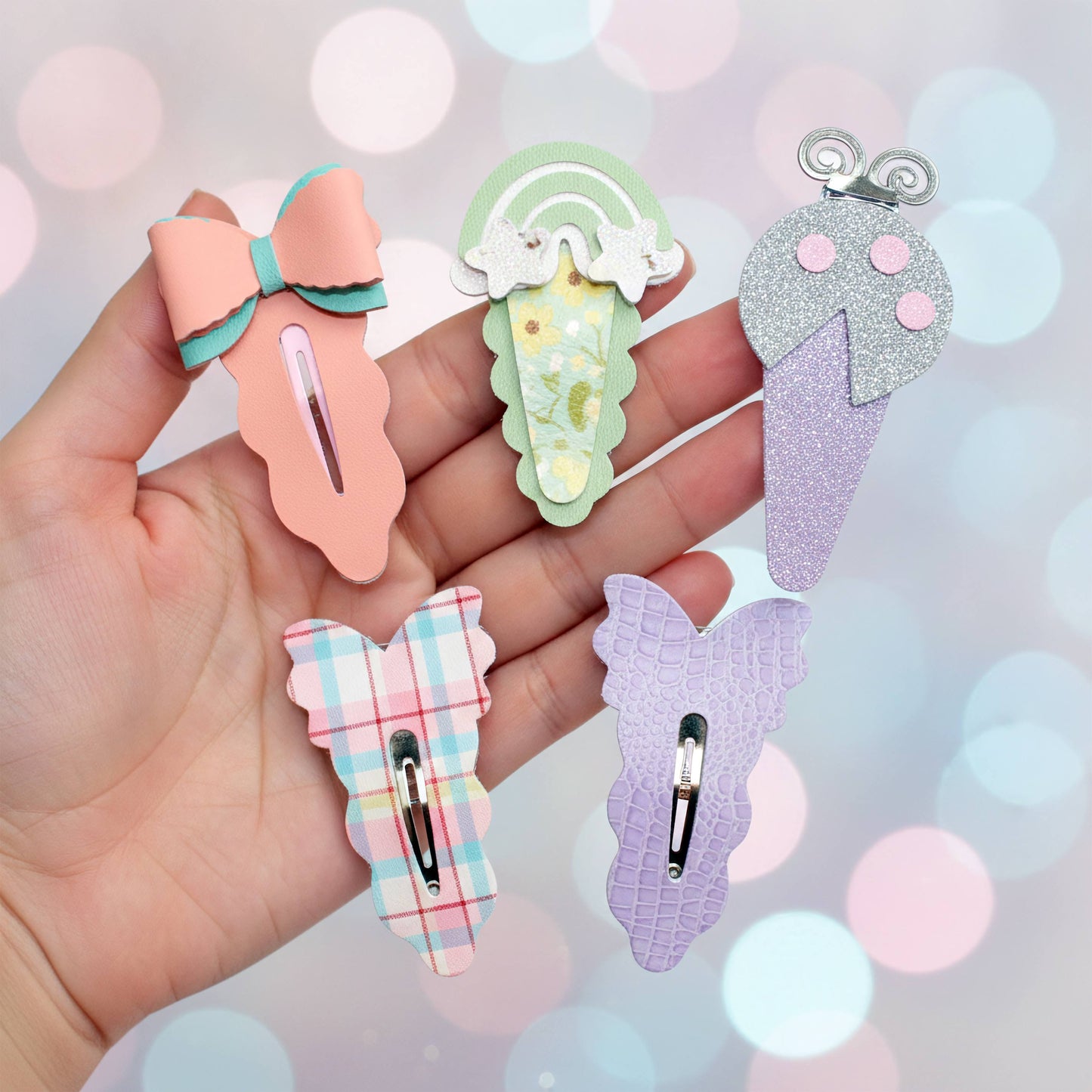 A hand holding five colorful hair clips, against a sparkling pastel background.Four cute hair clips held in a hand against a pastel bokeh background. The clips feature pastel designs including a peach bow with mint green accents, a mint green ice cream cone with white swirls, a glittery lavender ice cream cone with pink scoops, and a lavender plaid bow with pink and blue checks.