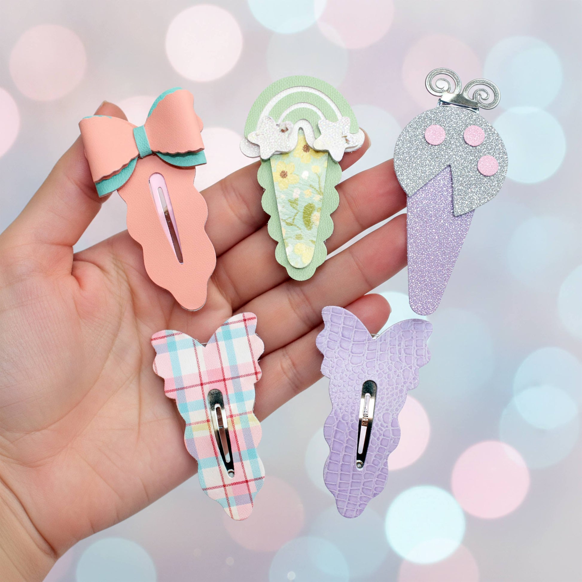 A hand holding five colorful hair clips, against a sparkling pastel background.Four cute hair clips held in a hand against a pastel bokeh background. The clips feature pastel designs including a peach bow with mint green accents, a mint green ice cream cone with white swirls, a glittery lavender ice cream cone with pink scoops, and a lavender plaid bow with pink and blue checks.