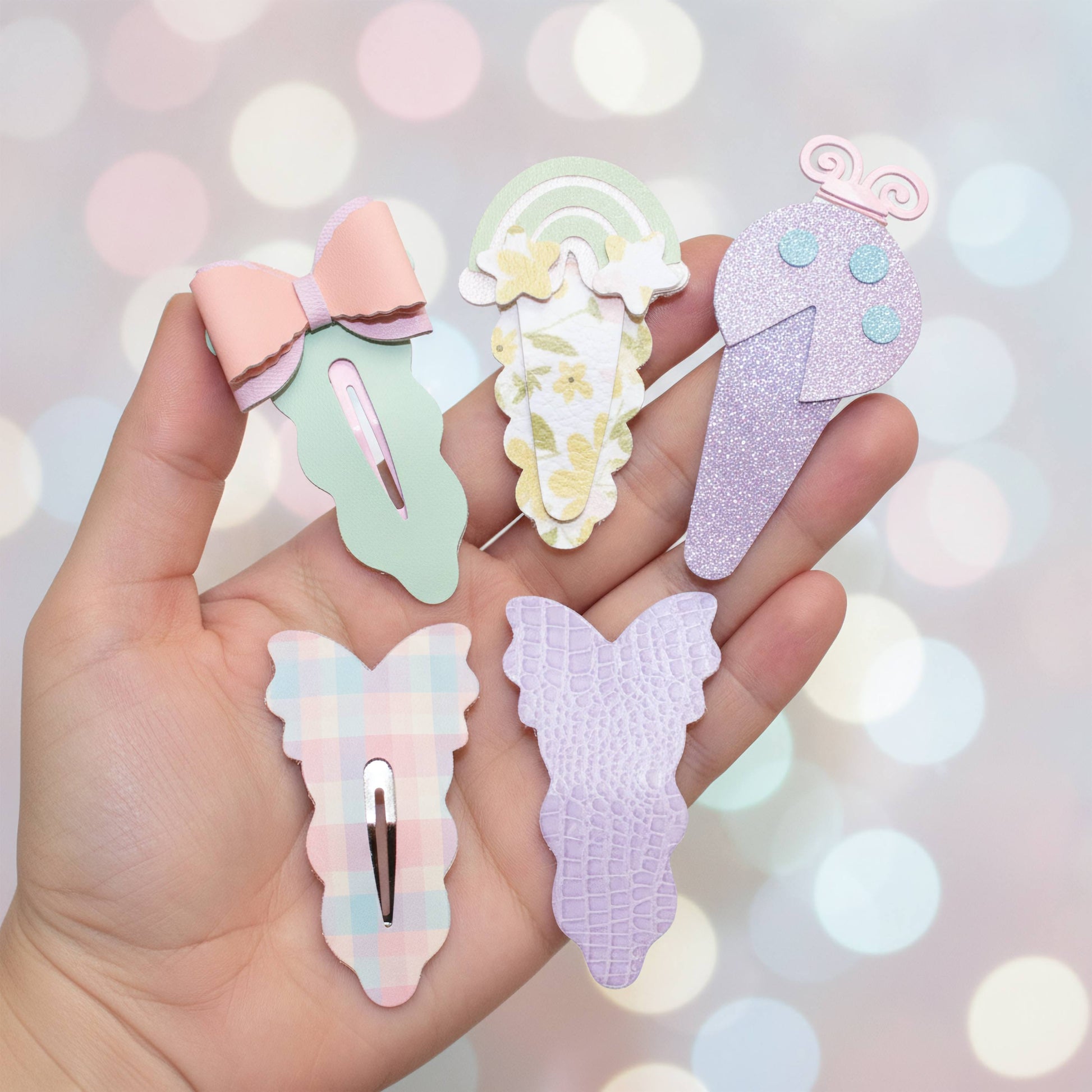 A hand holds five pastel hair clips shaped like a bow, a rainbow ice cream cone, a glitter ice cream cone, a plaid bunny, and a crocodile texture bunny against a soft pastel bokeh background.