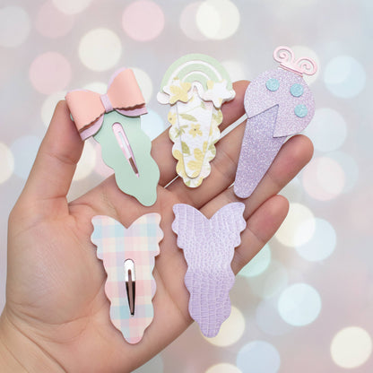 A hand holds five pastel hair clips shaped like a bow, a rainbow ice cream cone, a glitter ice cream cone, a plaid bunny, and a crocodile texture bunny against a soft pastel bokeh background.