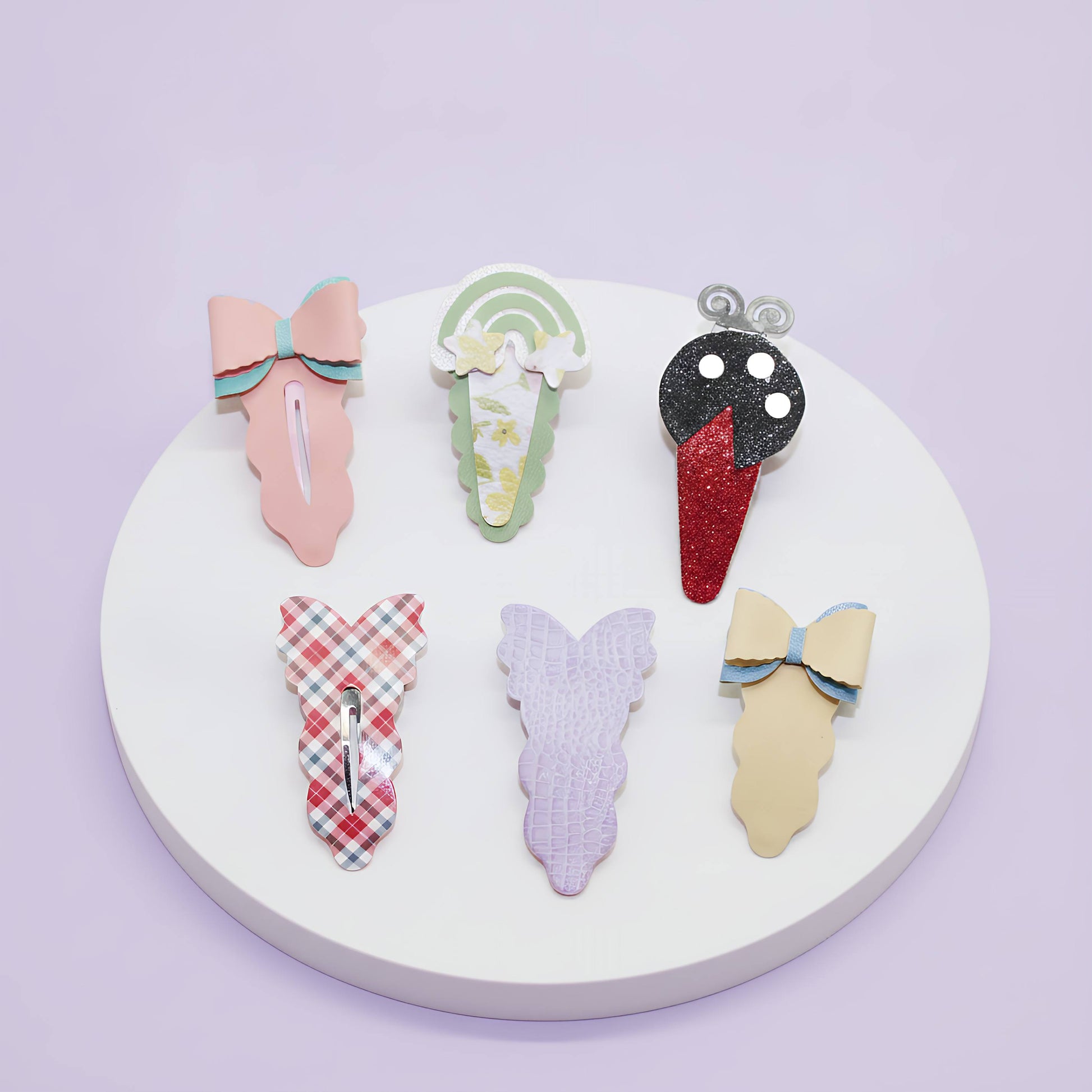 Six decorative hair claw clips arranged on a round white display plate against a pale lavender background. The clips include pastel pink, mint green, black-and-red glitter, red-and-white plaid, lavender, and cream with blue trim.