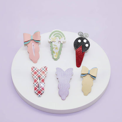 Six decorative hair claw clips arranged on a round white display plate against a pale lavender background. The clips include pastel pink, mint green, black-and-red glitter, red-and-white plaid, lavender, and cream with blue trim.