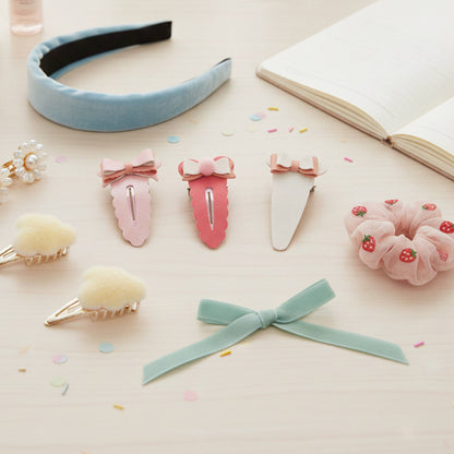 A styled flat-lay of pastel hair accessories on a light wood surface, including a light blue velvet headband, three pink bow hair clips, a pale pink scrunchie with strawberry details, fluffy cream hair clips, and a mint green ribbon bow, with an open book in the background.