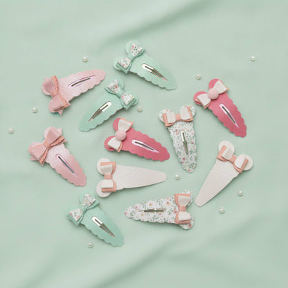 A set of pastel hair claw clips arranged on a mint green fabric background with small pearl-like beads. The clips come in pink, mint green, white, and floral print, each decorated with a small bow and ribbon accent.