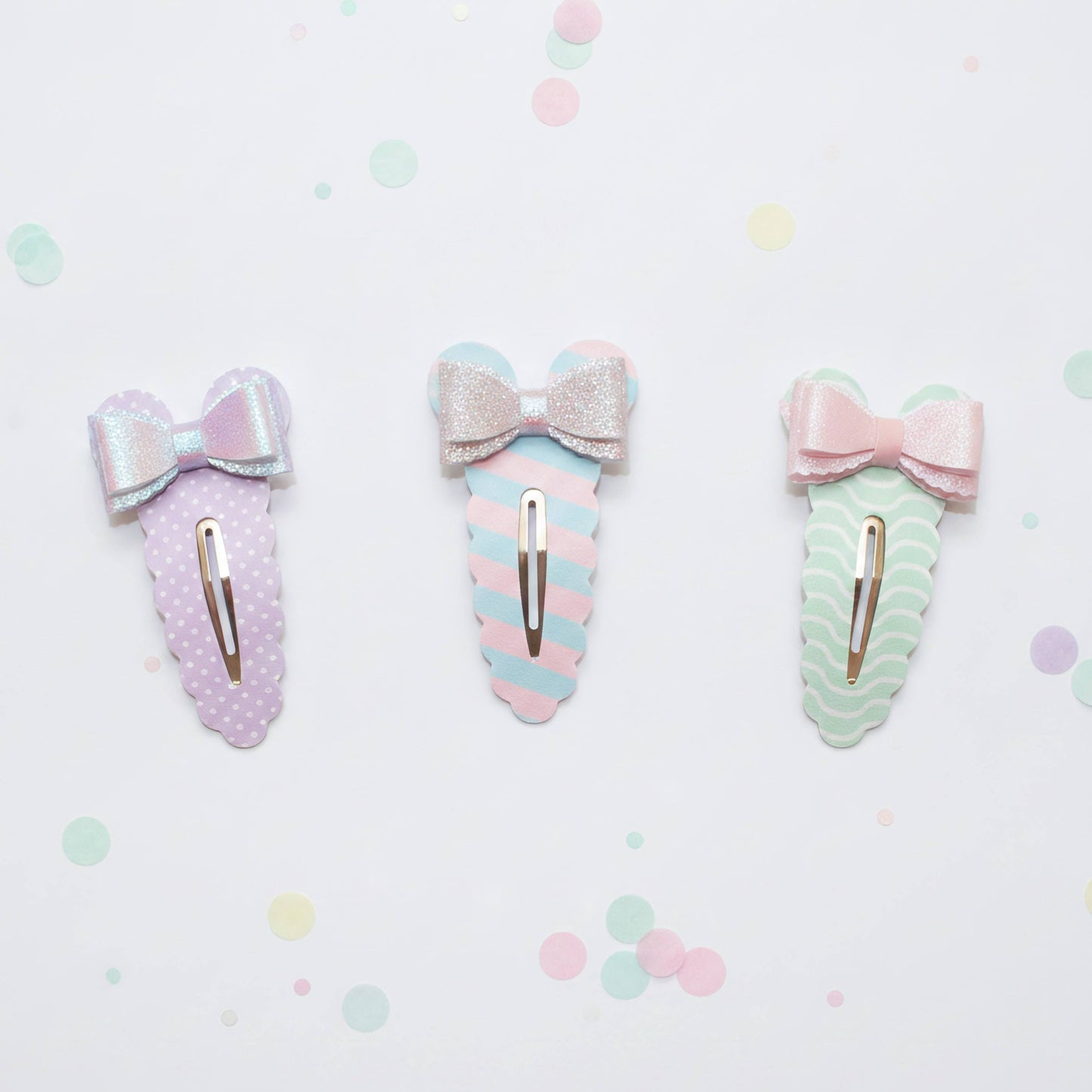 Three pastel hair claw clips with bow details, arranged horizontally on a white background with scattered pastel confetti dots. The left clip is lavender with white polka dots and a silver glitter bow.