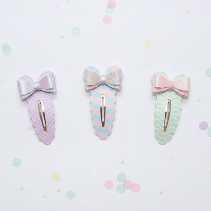 Three pastel hair claw clips with bow details, arranged horizontally on a white background with scattered pastel confetti dots. The left clip is lavender with white polka dots and a silver glitter bow.