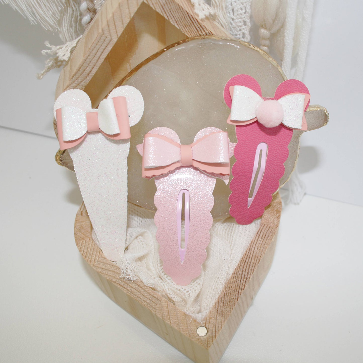 Three pastel hair clips with bow designs displayed in a small wooden box lined with cream fabric. The clips are white, light pink, and medium pink, each featuring a bow detail in a matching or contrasting pastel color.