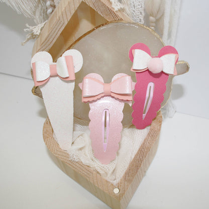 Three pastel hair clips with bow designs displayed in a small wooden box lined with cream fabric. The clips are white, light pink, and medium pink, each featuring a bow detail in a matching or contrasting pastel color.