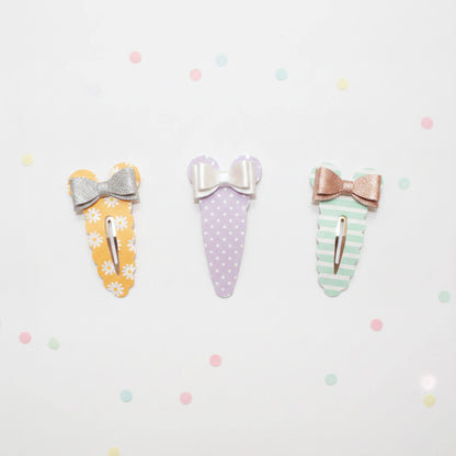 Three cute bow hair clips arranged in a row on a white background with pastel confetti. The left clip is yellow with white daisy patterns and a silver glitter bow.