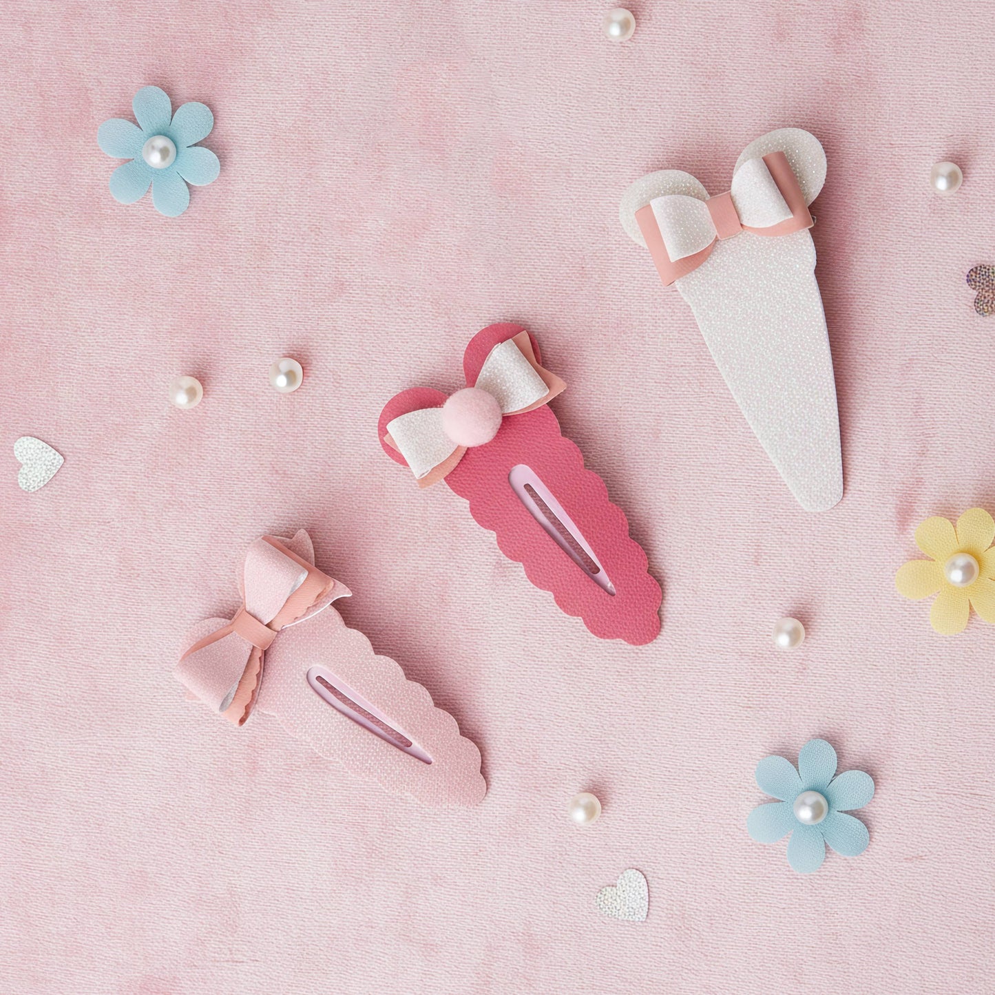 Three pastel-colored hair clips arranged on a pink textured background with decorative flowers, pearls, and small hearts. The clips are white, light pink, and dark pink, each adorned with a matching bow and pearl accent.