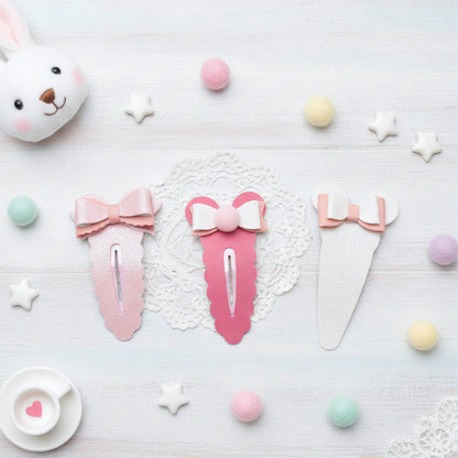 Three pastel bow hair clips arranged on a white textured surface with decorative props, including a bunny plush, pastel pom-poms, star-shaped candies, lace, and a small teacup.