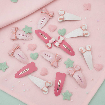 A set of pastel pink and white hair clips arranged on a soft pink fabric background with small decorative hearts, stars, and pearls.