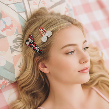 A blonde woman lying down wearing three decorative hair clips, including a red-and-white striped clip with white oval details and two black-and-red patterned clips, against a pastel patterned pillow and pink check background.