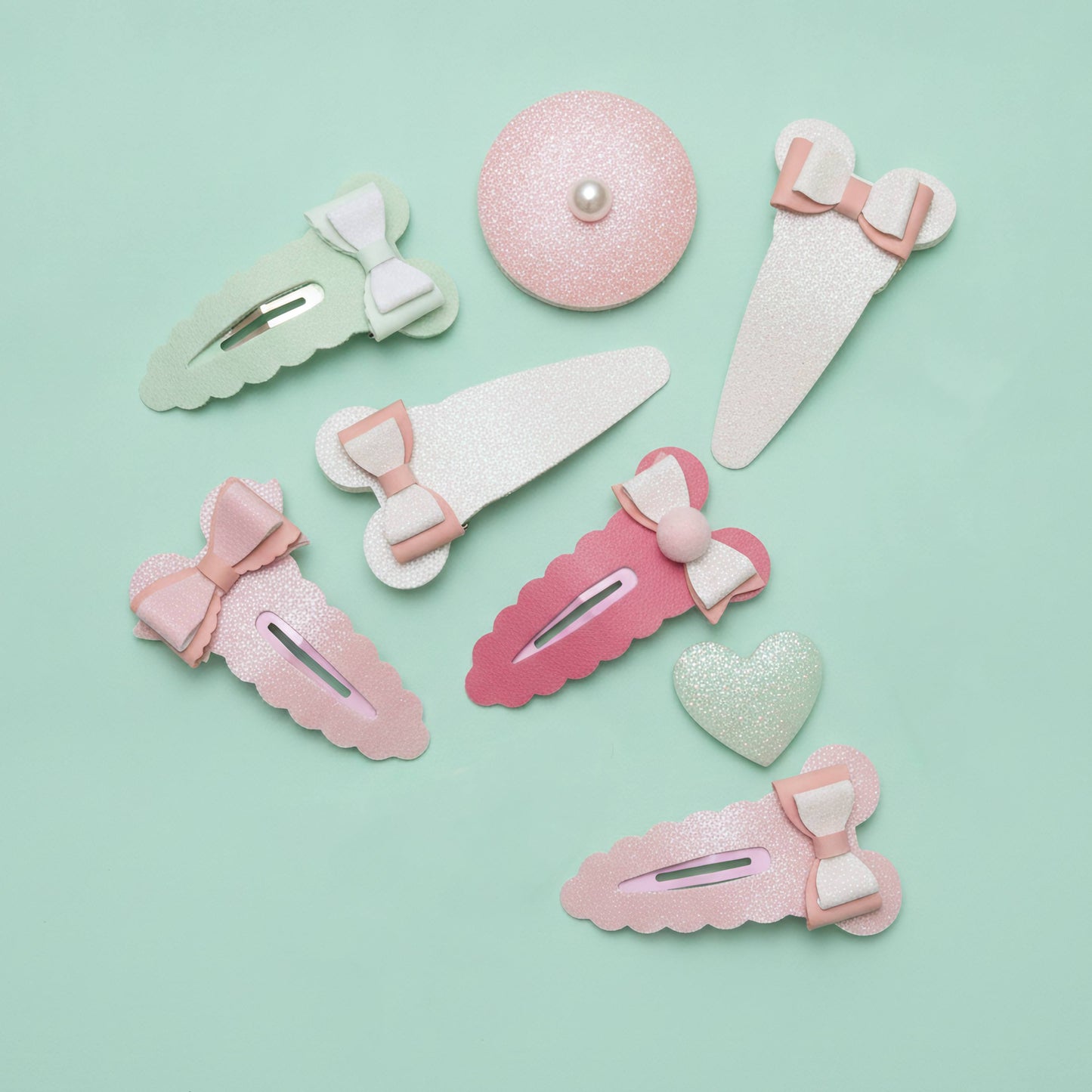 A set of pastel hair clips and barrettes arranged on a mint green background. The set includes various shapes like bows, hearts, and scalloped edges in pink, light green, and white glitter, with one round clip featuring a small pearl center.