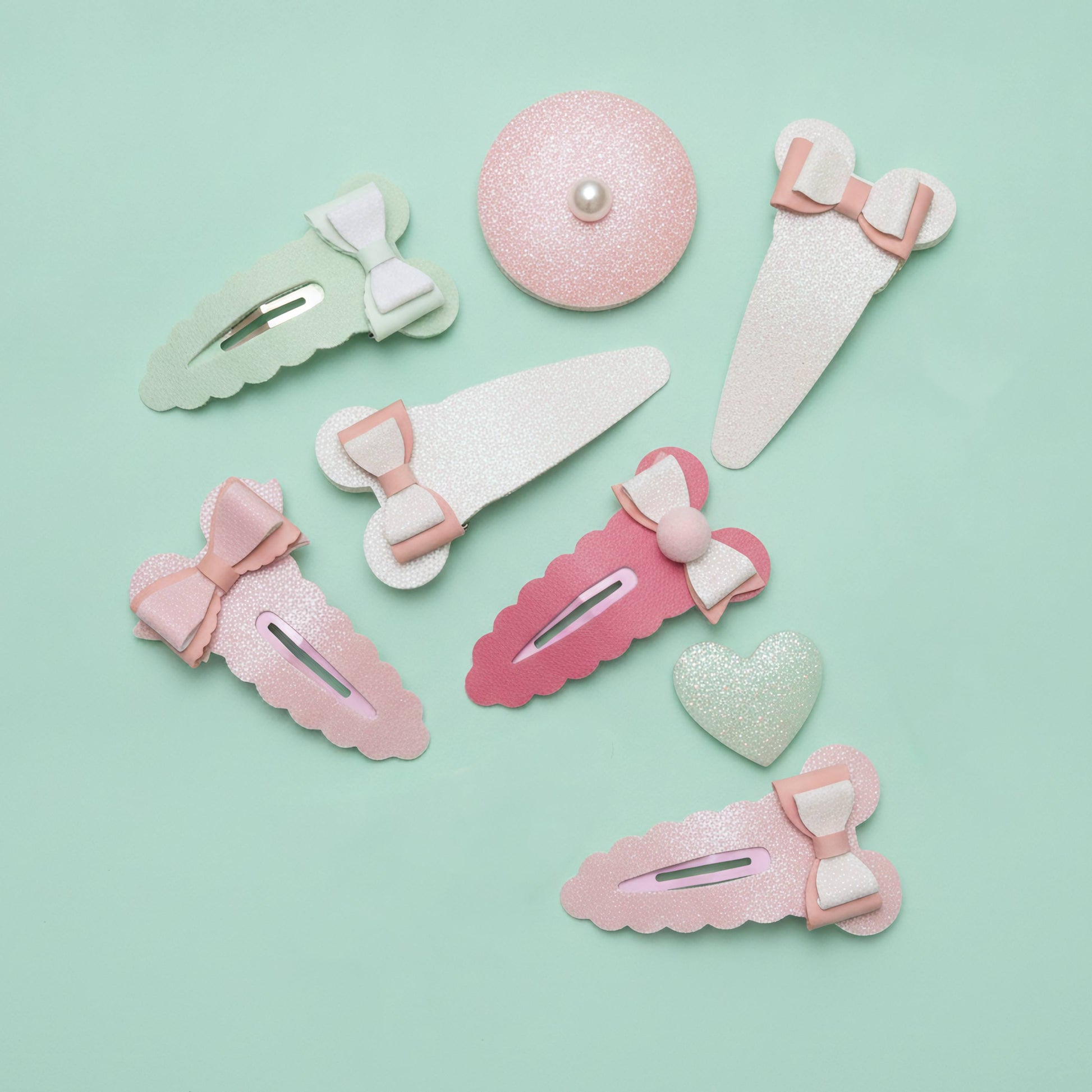 A set of pastel hair clips and barrettes arranged on a mint green background. The set includes various shapes like bows, hearts, and scalloped edges in pink, light green, and white glitter, with one round clip featuring a small pearl center.