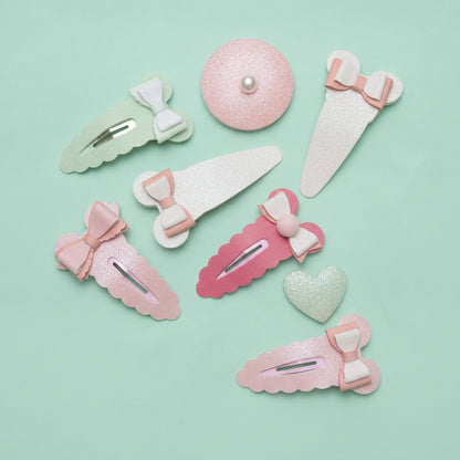 A set of pastel hair clips and barrettes arranged on a mint green background. The set includes various shapes like bows, hearts, and scalloped edges in pink, light green, and white glitter, with one round clip featuring a small pearl center.