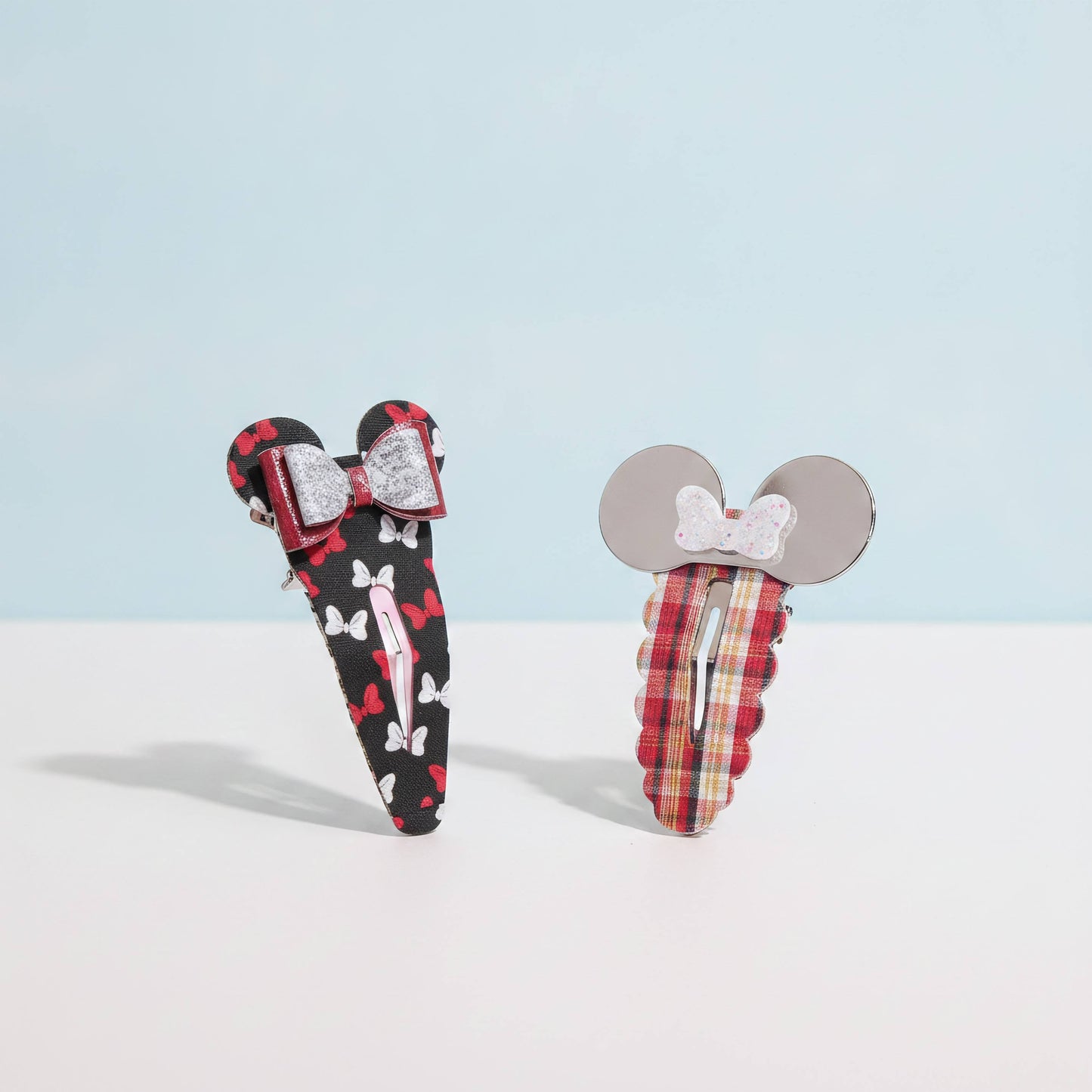 Two fabric hair clips styled like girl  Mouse inspired bows and ears, standing upright on a white surface against a pale blue background. One clip is black with red and white motifs and a red bow; the other is red plaid with gray ears and a white bow.