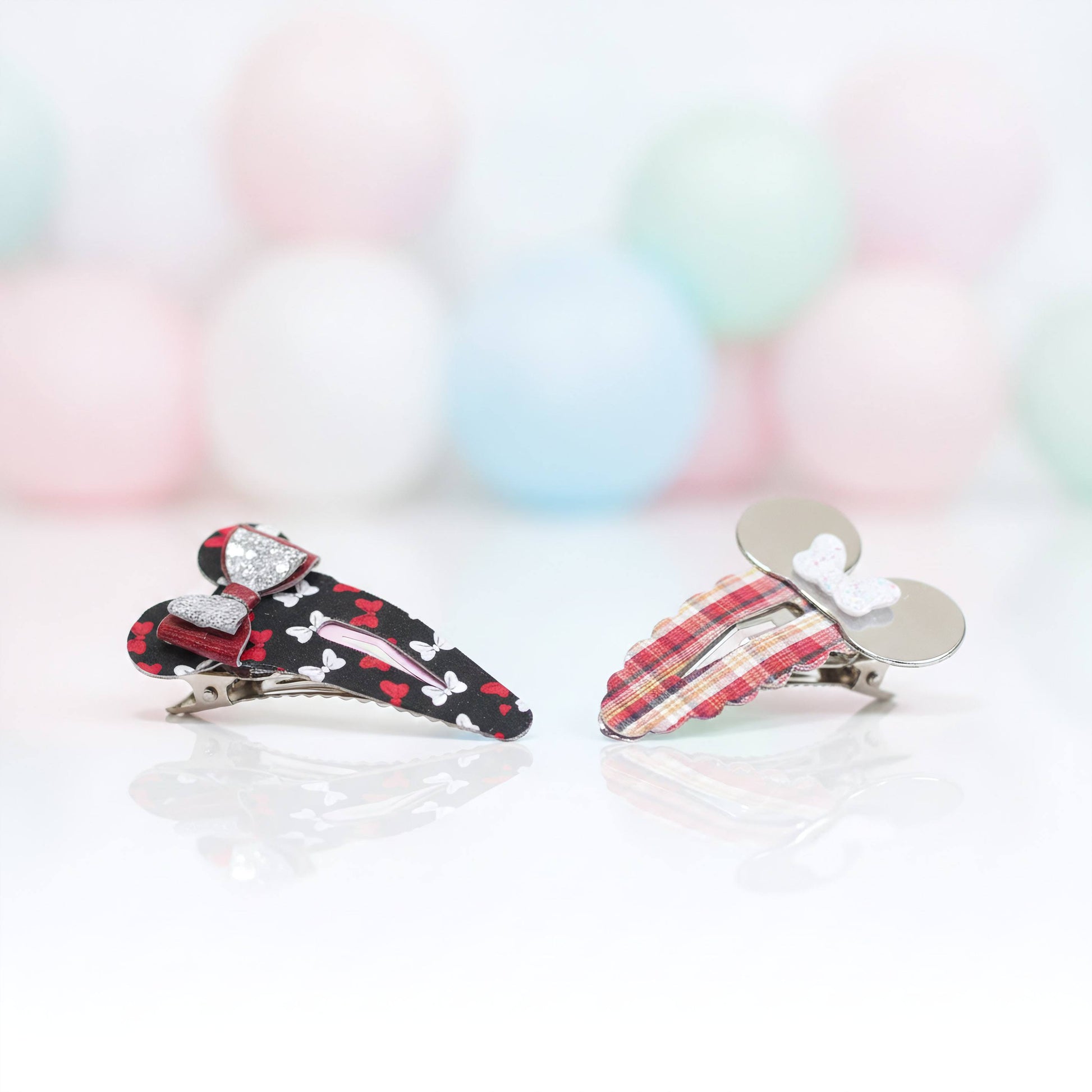 Two small hair claw clips on a white reflective surface with a soft pastel balloon background. One clip is black with white bones, red hearts, and a red bow with rhinestones; the other is red plaid with a white bow and silver metal ears.