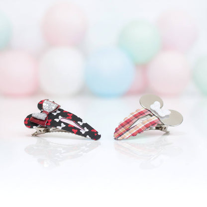 Two small hair claw clips on a white reflective surface with a soft pastel balloon background. One clip is black with white bones, red hearts, and a red bow with rhinestones; the other is red plaid with a white bow and silver metal ears.