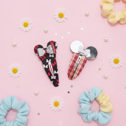 Two hair clips styled like Mouse ears with bows, displayed on a pink background with daisy decorations and scrunchies. One clip has a black and red pattern with white bows, and the other has a red and yellow plaid with a silver bow.