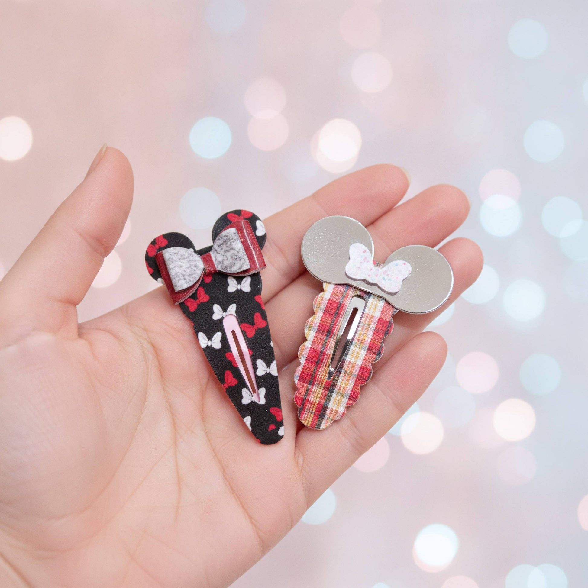 Two decorative hair clips shaped like mouse ears are held in a person's hand against a soft pink bokeh background. One clip is black with red and white bow patterns and a silver bow; the other is silver with a red plaid pattern and a white bow accent.