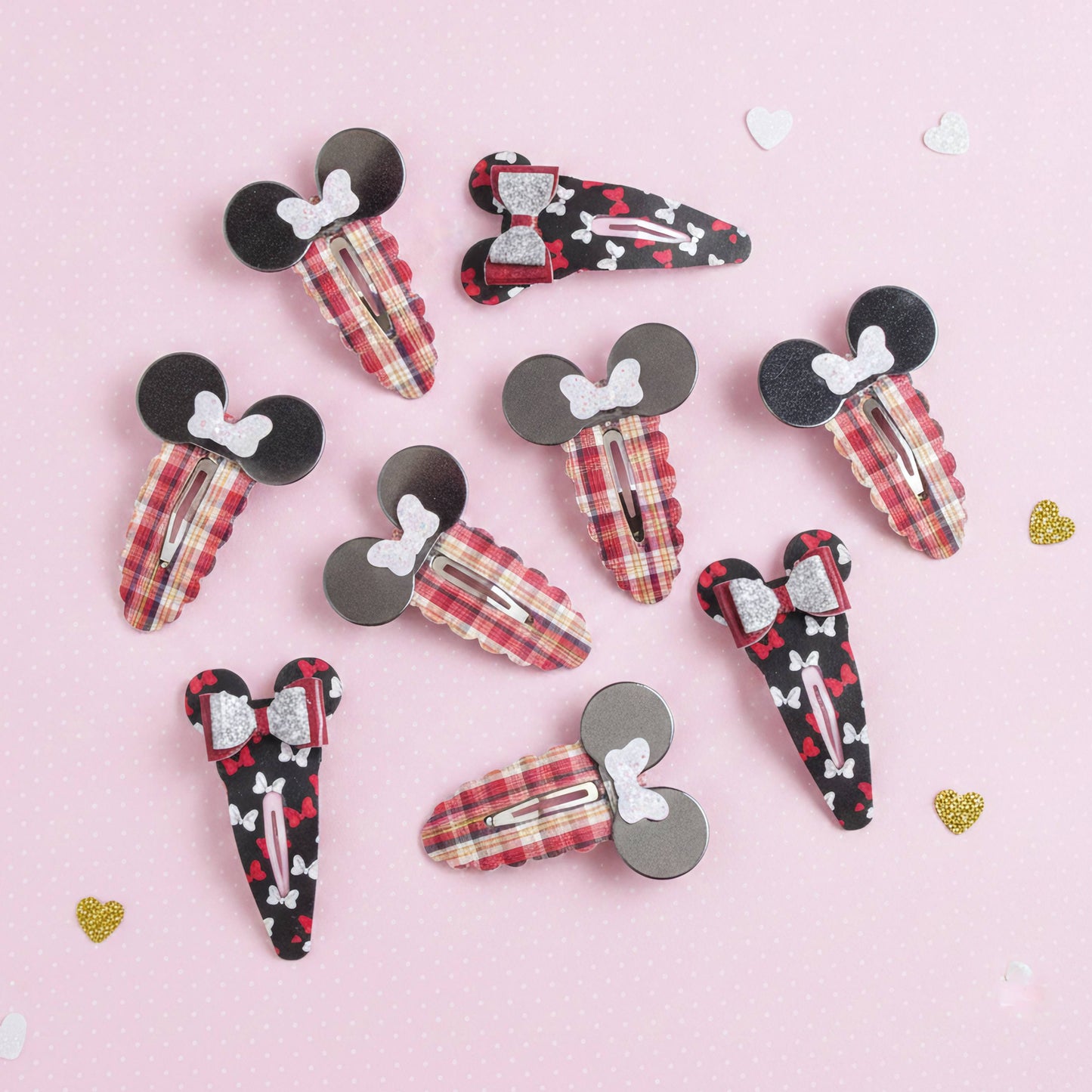 A set of ten hair claw clips styled like Mouse and Minnie Mouse silhouettes, arranged on a light pink background with small scattered heart decorations. The clips feature various fabric patterns including red and beige plaid, black and red hearts, and black and white stripes, with some clips featuring glittery silver bows.