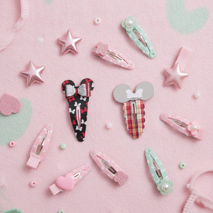 A styled flat-lay of multiple pastel mouse hair clips arranged on a pink textured background with mint green heart shapes and small pearl-like beads. The clips include pink, mint green, red plaid, black-and-white patterned, and silver-gray designs, with some featuring bow details and star or heart accents.