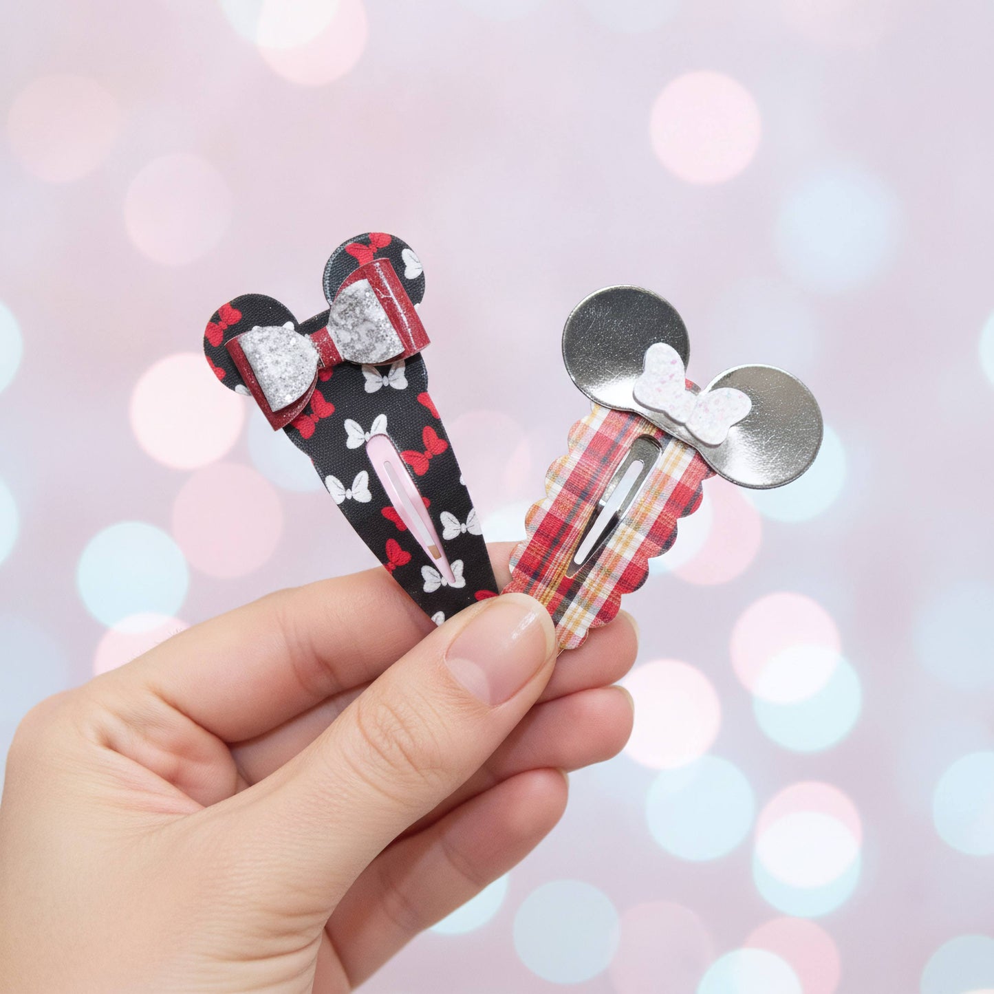 A hand holding two Mouse-themed hair clips against a soft pink bokeh background. One clip is black with white bows and a red bow detail, and the other is red and beige plaid with a white bow and silver mouse ears.