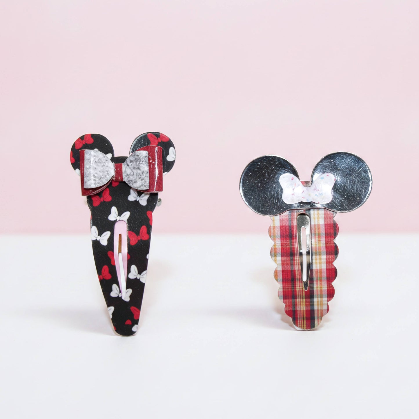 Two decorative hair clips shaped like Mouse ears, shown on a clean white and pale pink background. One clip has a black base with red bows and white butterflies, and the other has a red plaid base with a white bow.