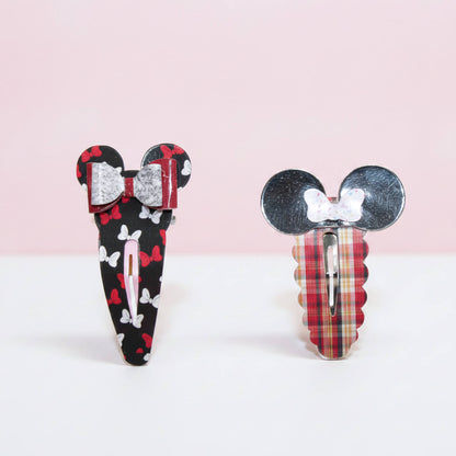 Two decorative hair clips shaped like Mouse ears, shown on a clean white and pale pink background. One clip has a black base with red bows and white butterflies, and the other has a red plaid base with a white bow.