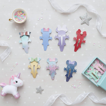 A styled flat-lay of pastel mouse-shaped hair clips arranged on a white polka-dot surface with lace ribbon, a cup of confetti, a small unicorn toy, and an open box of matching clips. The clips come in multiple pastel colors including blue, purple, red, yellow, pink, and glitter finishes.