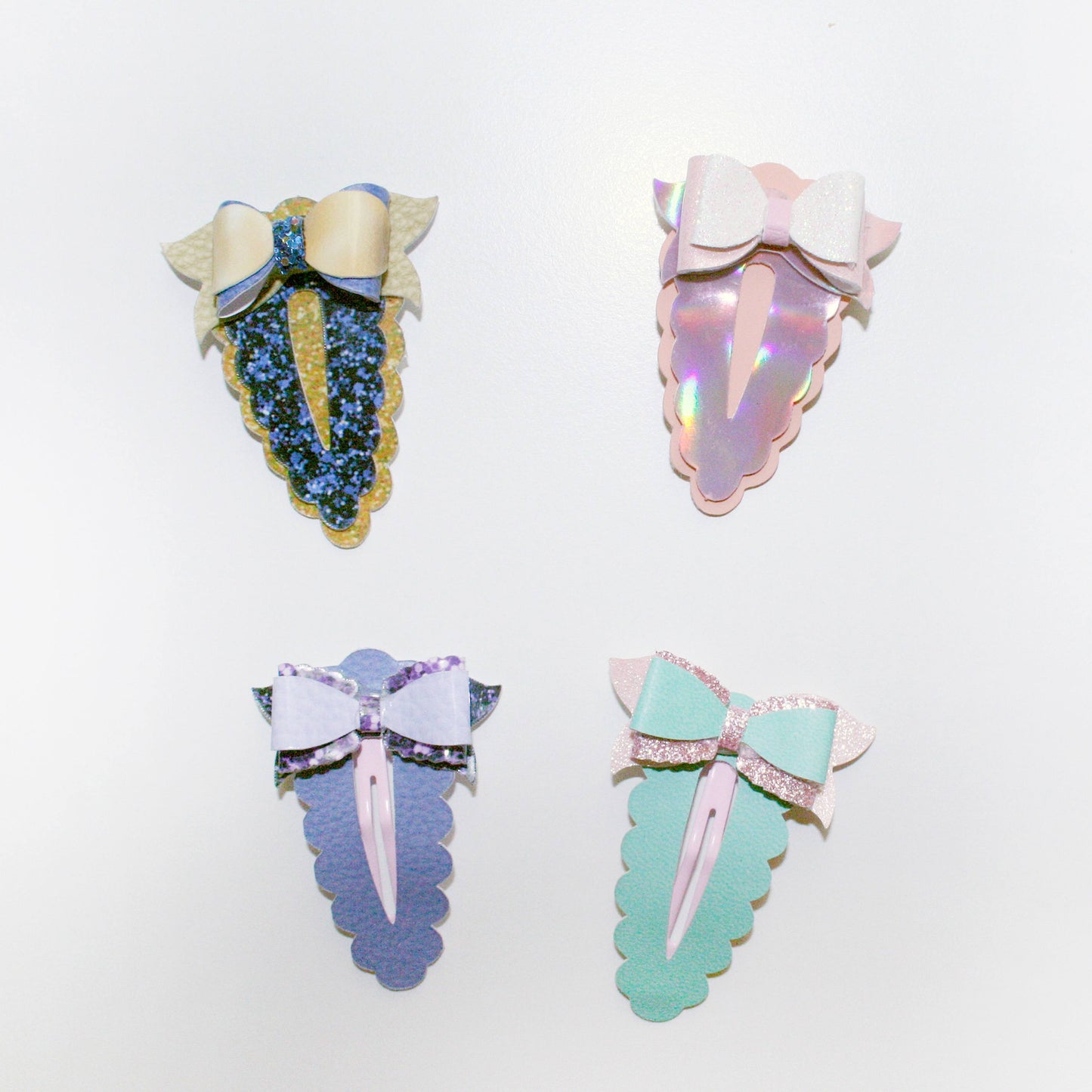 Four decorative hair claw clips arranged  on a plain white background. Each clip has a bow and iridescent scalloped body in pastel colors: blue and gold, pink and iridescent, lavender and black, and mint green and pink.