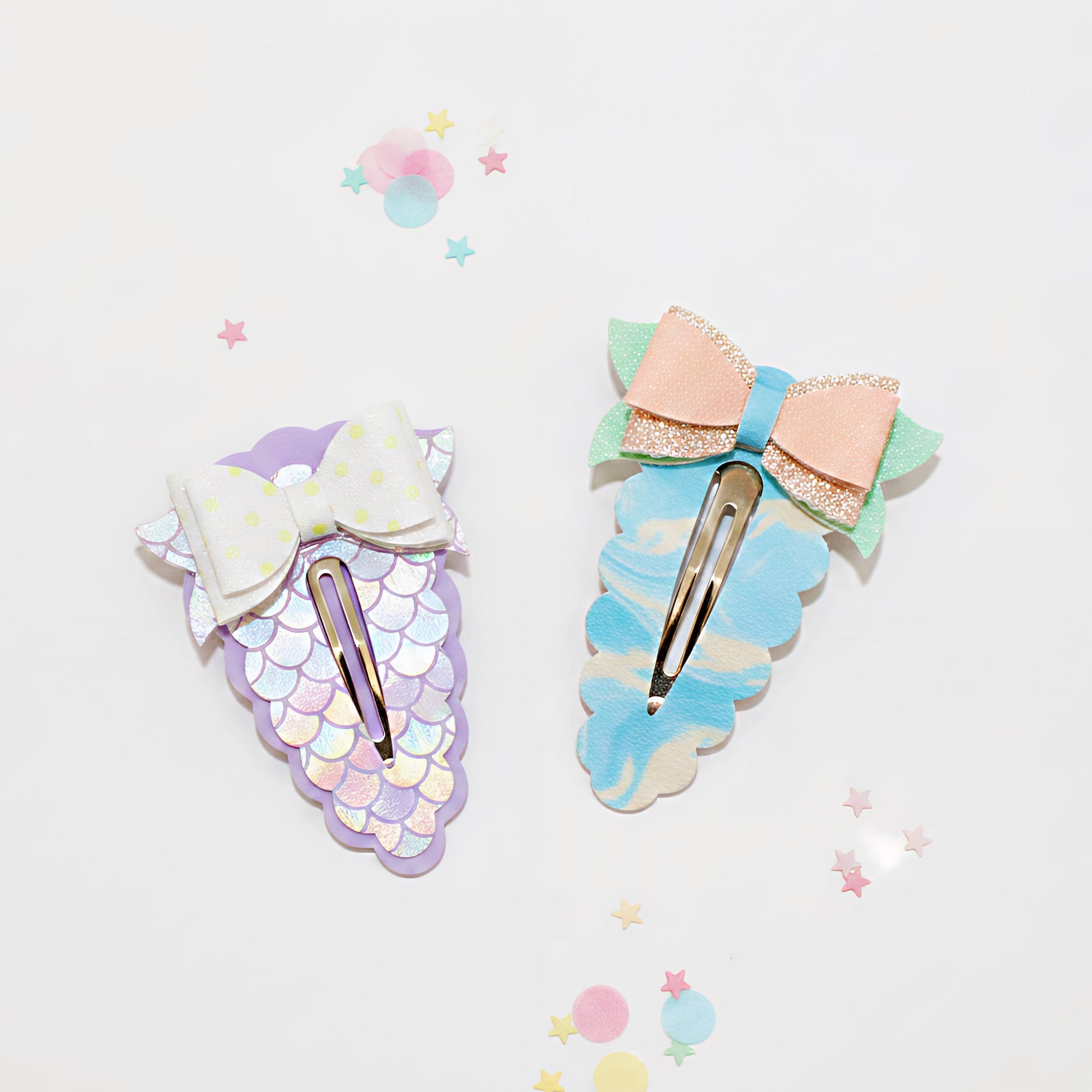 Two mermaid-themed hair clips on a white background with pastel star confetti. One clip is lavender with a white bow and pastel scale pattern, and the other is light blue with a peach glitter bow and mint green leaf accents.