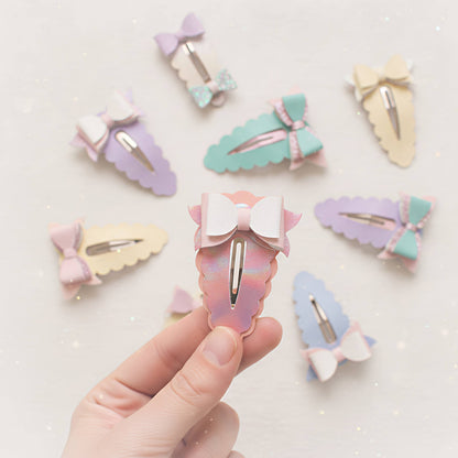 A hand holds a pastel iridescent hair clip with a white bow, surrounded by several other pastel-colored bow hair clips on a light sparkly background.