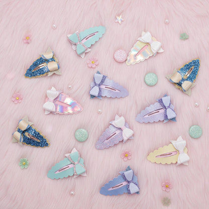 A flat-lay arrangement of pastel hair clips on a pink faux fur background. The clips feature pastel shades of blue, purple, mint green, and yellow, some with glittery finishes, iridescent holographic accents, and white bows.
