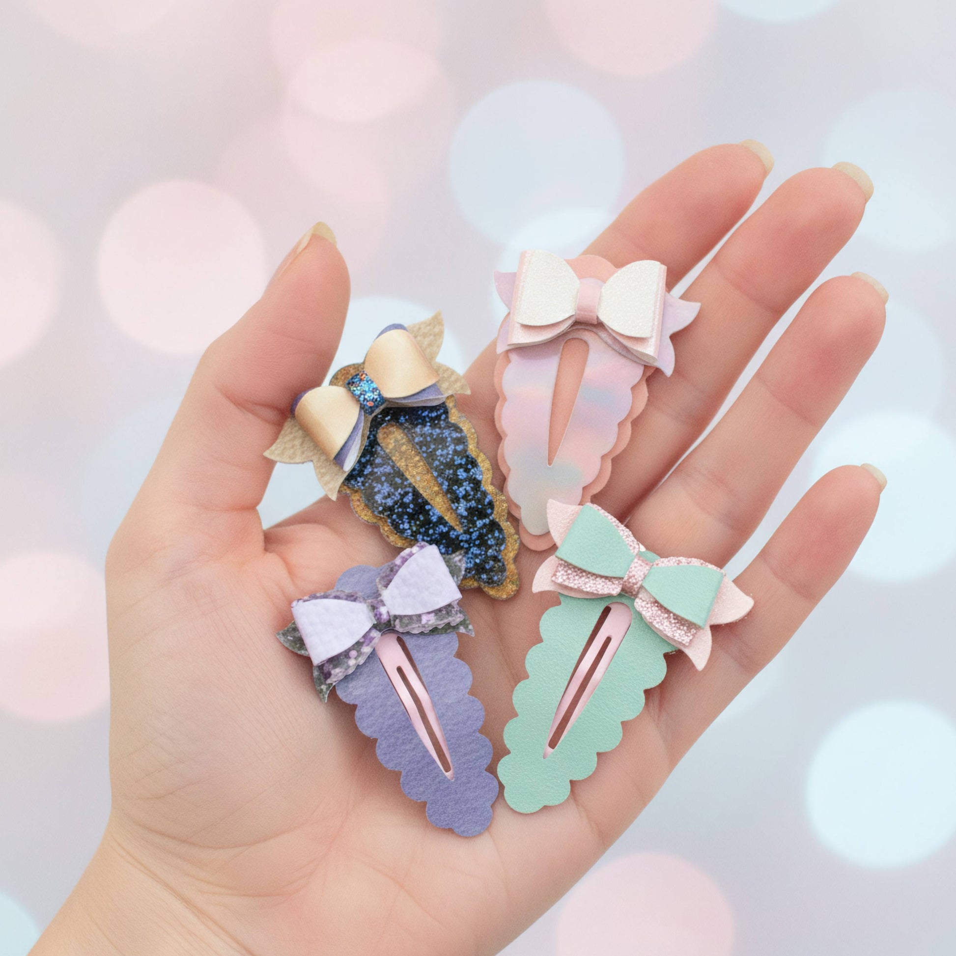 Four pastel hair claw clips held in a hand against a soft pink bokeh background. The clips feature glittery and satin bow details in lavender, mint green, light pink, and navy blue.