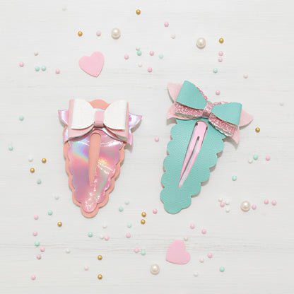 Two pastel cute hair clips styled on a white wooden background with scattered decorative hearts, pearls, and confetti. One clip is iridescent pink with a white bow, and the other is mint green with a pink and white bow.